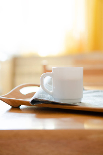 Λευκή κούπα καφέ με χαρτοπετσέτα στο τραπέζι σε πραγματικό εσωτερικό κουζίνα με έντονο φυσικό φως του ήλιου - Φωτογραφία, εικόνα
