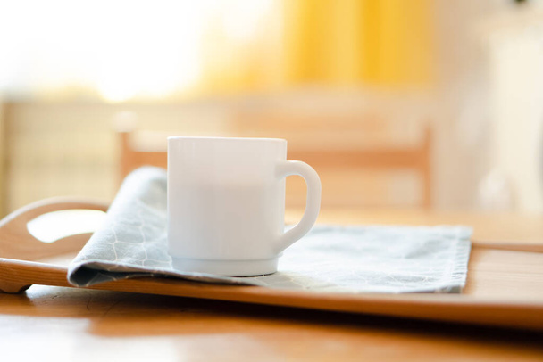 Witte koffiemok met servet op tafel in echt keukeninterieur met helder natuurlijk zonlicht - Foto, afbeelding