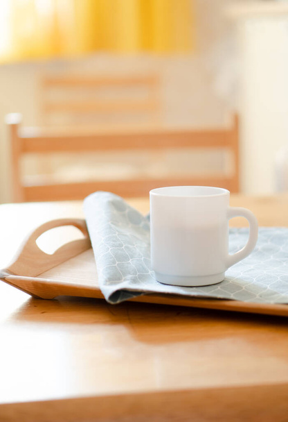 Witte koffiemok met servet op tafel in echt keukeninterieur met helder natuurlijk zonlicht - Foto, afbeelding