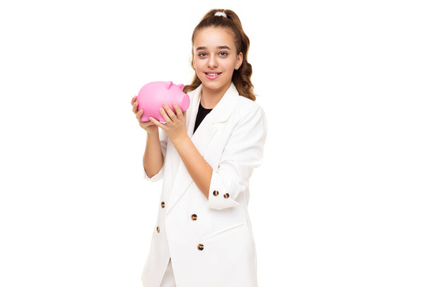 Foto van mooie schattige positieve glimlach brunette tienermeisje met paardenstaart in stijlvolle witte jas en witte broek met roze penny varken voor munten geïsoleerd op witte achtergrond met kopie - Foto, afbeelding