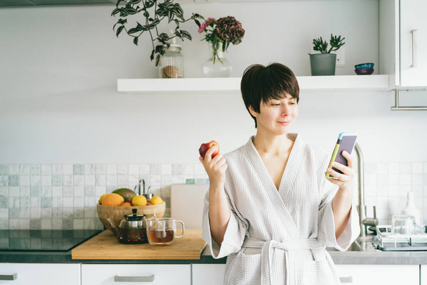 Χαμογελαστή γυναίκα με τη ρόμπα κοιτάζει το κινητό της τηλέφωνο και τρώει ένα μήλο ενώ στέκεται στην κουζίνα του Μόρντερν. Πρωινές συνήθειες και τελετουργίες. Εθισμός στο διαδίκτυο, σχεδιασμός ημέρας. - Φωτογραφία, εικόνα