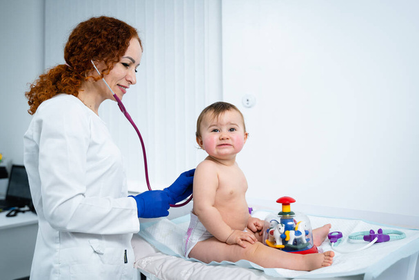 Pediatrician doctor concept. Children medical care. One year old baby girl examined by female pediatrician in clinic office. Child visiting doctor for health check-up. Doctor examine little patient. - Photo, Image