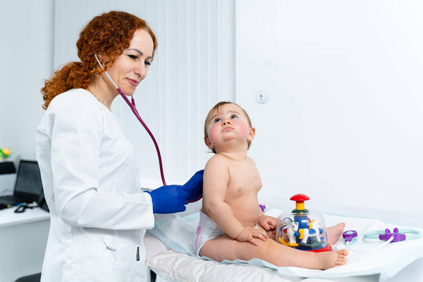 Red-haired female doctor examines little child for less than a year in a modern clinic. Pediatrician during examination baby in the hospital. Childrens healthcare. Diagnostics and treatment for kid. - Photo, Image