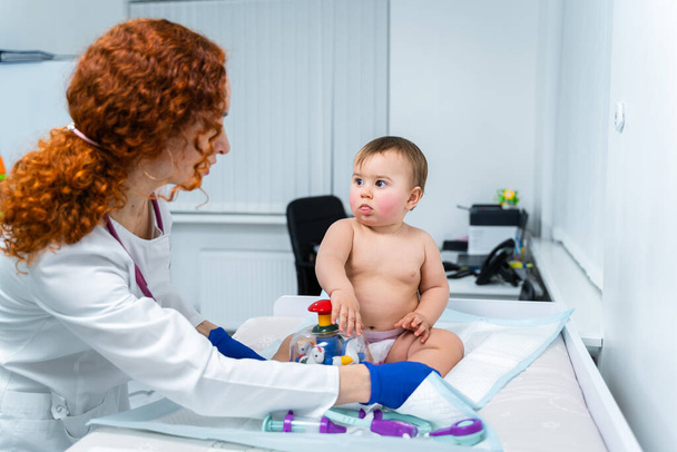 La doctora pelirroja examina a un niño pequeño por menos de un año en una clínica moderna. Pediatra durante el examen del bebé en el hospital. Salud infantil. Diagnósticos y tratamiento para niños. - Foto, Imagen