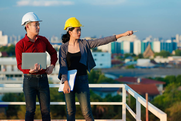 Занятые азиатские инженеры мужского и женского пола в команде хардхатов, работающие вместе, стоя на крыше и работая над проектом, указывая в сторону, открытый снимок с голубым небом. - Фото, изображение