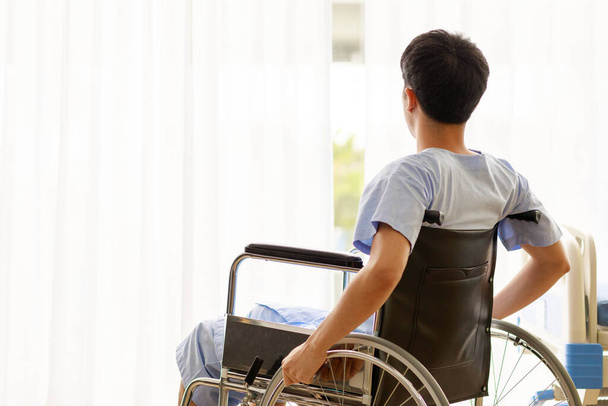 Asian male hopeful patient in blue hospital uniform sit in wheelchair look outside through white clear curtain of ward room in warm light while has hope to recover from serious injury legs problem. - 写真・画像