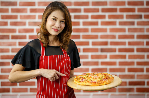 Asiatische schöne Frau trägt rote Schürze, lächelt und zeigt mit dem Finger auf hausgemachte Pizza mit Schinken, Paprika auf hölzernem Tablett, steht in der Küche mit Backsteinmauer mit leerem Kopierraum - Foto, Bild
