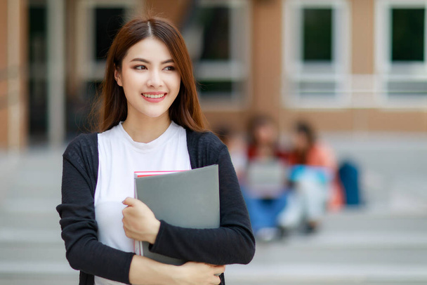 Young and beautiful Asian college student girls holding books, pose to camera with group of friends blur in background against school building. Learning and friendship of teens close friend concept. - Photo, Image