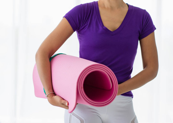 Asian beautiful sportive healthy woman with wearing purple shirt and white pants, posing and smiling with confidence and happiness while holding pink yoga mat and looking to the camera - Photo, Image