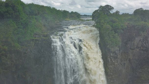 Řeka Zambezi teče podél skalnatého koryta a zřítí se do rokle. Zelená vegetace na březích. Mlha nad propastí. Victoria Falls. Zimbabwe. - Fotografie, Obrázek