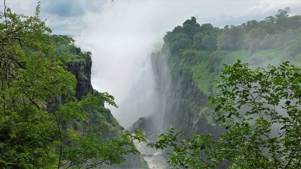 Il y a un épais brouillard de Victoria Falls pulvérisation sur la gorge. Une rivière orageuse coule au fond du canyon. Il y a une végétation verte autour. Zimbabwe - Photo, image
