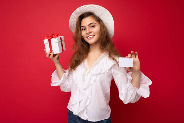 Portrét pozitivní veselý módní žena ve formalwear držení dárkové krabice a kreditní karty při pohledu na kameru izolované na červeném pozadí s kopírovacím prostorem - Fotografie, Obrázek