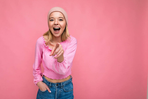 Photo de jeune positif heureux étonné belle femme blonde avec des émotions sincères portant chemisier rose et chapeau rose isolé sur fond rose avec l'espace de copie et pointant du doigt à la caméra - Photo, image