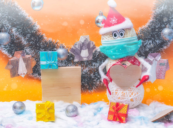 Le bonhomme de neige du Nouvel An en tenue rouge et masque de protection sur son visage se dresse sur un fond de fête lumineux. Il tient un cœur en bois propre dans ses mains. - Photo, image
