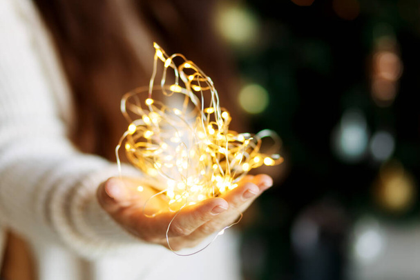 Christmas miracle - woman in white sweater holds glowing led light garland in hands, bokeh background - Photo, Image