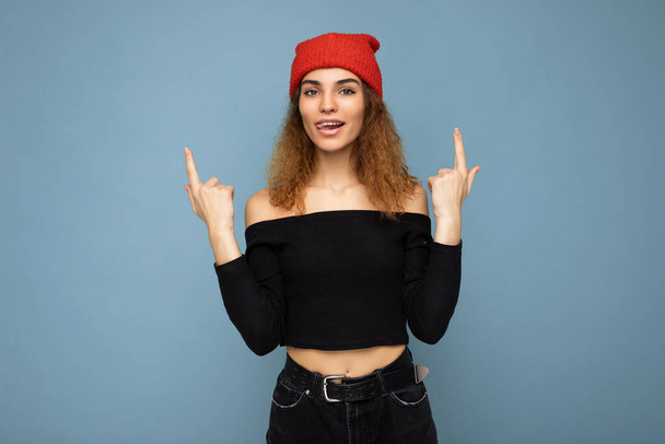 Photo of young positive happy attractive brunette curly woman with sincere emotions wearing black crop top and red hat isolated on blue background with empty space and pointing up at free space - Fotó, kép