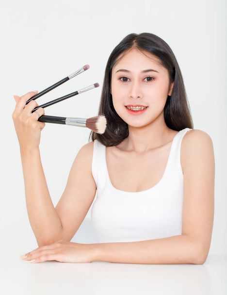 Portré szépség stúdió lövés ázsiai rövid fekete haj női modell fogszabályzóval tartály felső mellény fehérnemű tartsa három méret alapítvány kefék a kézben mosoly nézd meg a kamerát fehér háttér. - Fotó, kép