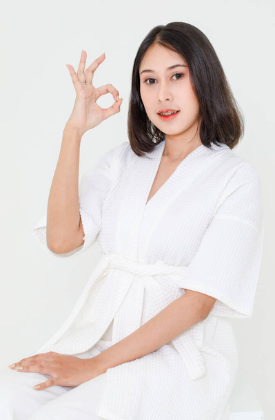 Beauty portrait studio shot of millennial young Asian black short hair female model wearing makeup in shower rope standing smiling look at camera posing holding ok hand sign up on white background. - 写真・画像