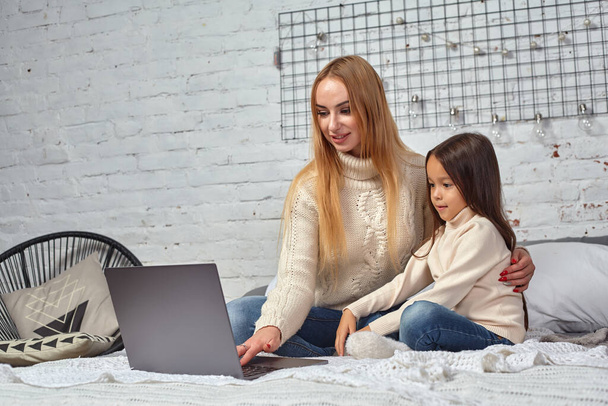 Schöne junge Mutter und ihre süße Tochter in weißen Pullovern und Jeans liegen zu Hause auf dem Bett, lachen und schauen in den Laptop - Foto, Bild