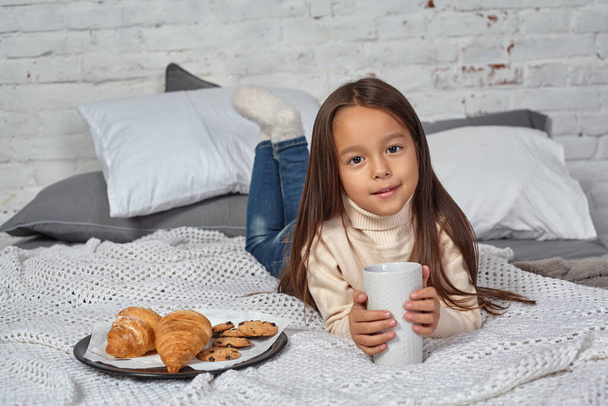 Csinos kislány, 6 éves, fehér pulóverben és farmerben. Gyerek a szobában egy ággyal, eszik croissant és tea- vagy kakaóivás. - Fotó, kép