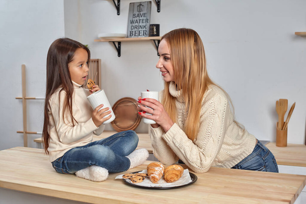 Shot of cheerful mother and daughter sit together at kitchen table, drink hot tea in morning, have pleasant friendly talk between each other. - Zdjęcie, obraz