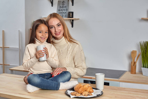 Shot of cheerful mother and daughter sit together at kitchen table, drink hot tea in morning, have pleasant friendly talk between each other. - Photo, Image