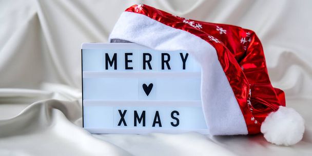 Lightbox con testo MERRY XMAS con cappello da Babbo Natale su fondo in tessuto di seta. Concetto vacanza invernale. Natale e Capodanno - Foto, immagini