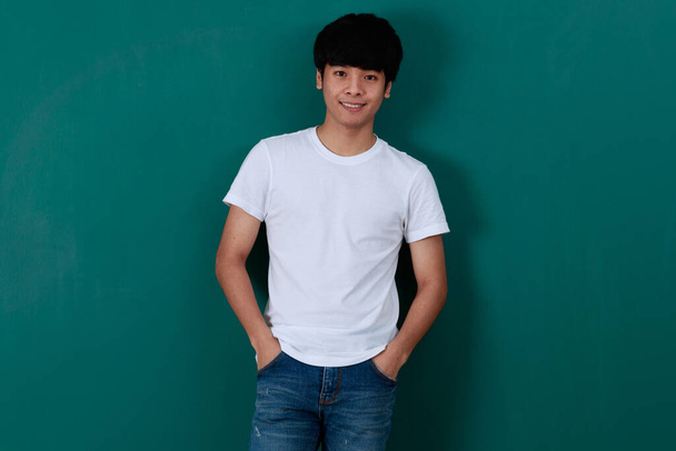 Deleitado joven asiático masculino en camiseta blanca y jeans de pie sobre fondo verde en el estudio y mirando a la cámara - Foto, Imagen