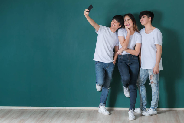 Group of cheerful Asian friends standing on green background in studio and taking self shot on smartphone - Fotoğraf, Görsel