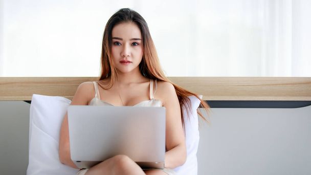 Young Asian woman with long hair wearing undergarment and looking at camera and using laptop while relaxing on bed in morning at home. - Foto, imagen