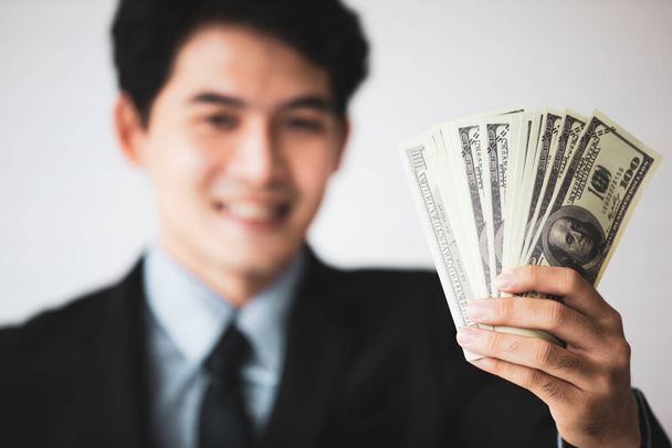 Selektiver Fokus auf Geld: Asiatische junge hübsche schwarze kurze Haare erfolgreicher reicher Geschäftsmann hält US-Dollar-Banknoten in der Hand im verschwommenen Vordergrund. - Foto, Bild