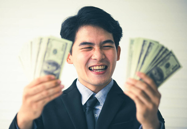 Close-up portret schot van Aziatische jonge knappe zwart kort haar succesvolle rijke zakenman draagt formele pak en stropdas lachen en voldoen met volledige US dollar honderd bankbiljetten munt in handen. - Foto, afbeelding