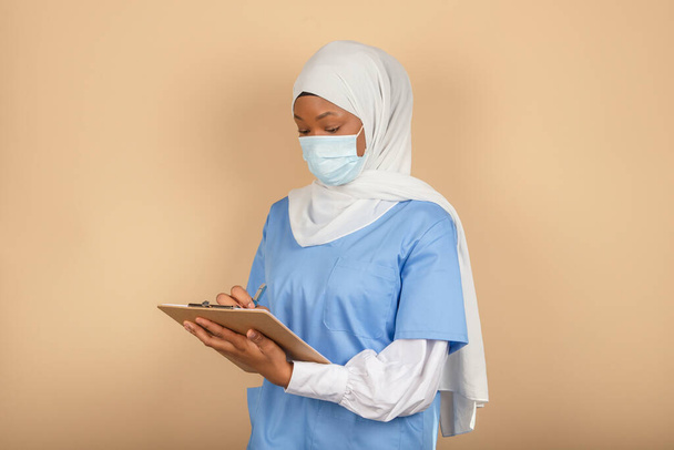 Una giovane infermiera musulmana in piedi con una maschera chirurgica prende nota su un blocco appunti su una parete gialla - Foto, immagini
