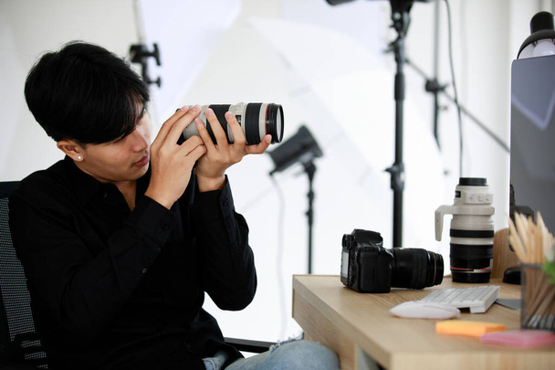 Asian young handsome happy male short black hair photographer hold and inspect big DSLR camera lens in hands sit on chair in photography studio - Фото, изображение