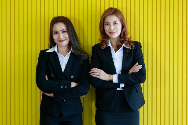 Selbstbewusste asiatische Geschäftsfrauen verschränken die Arme und lächeln in die Kamera, während sie sich im Büro an eine leuchtend gelbe Wand lehnen - Foto, Bild