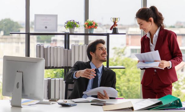 Pozytywny mężczyzna i kobieta w garniturach uśmiechnięci i dyskutujący o dokumentach podczas pracy nad projektem biznesowym we współczesnym miejscu pracy - Zdjęcie, obraz