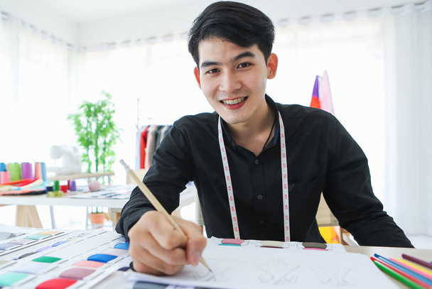 Content ethnic male clothes designer sitting at table with fabric samples and sketches with happy face. - Photo, Image
