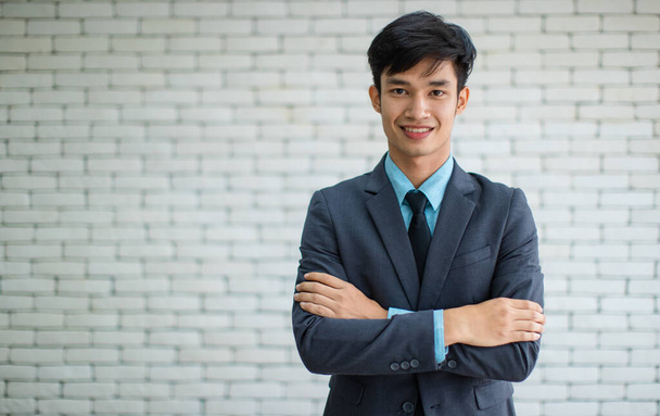 Šťastný mladý asijský manažer v obleku při pohledu na kameru s úsměvem a zkřížené paže na rozmazané pozadí cihlové zdi - Fotografie, Obrázek
