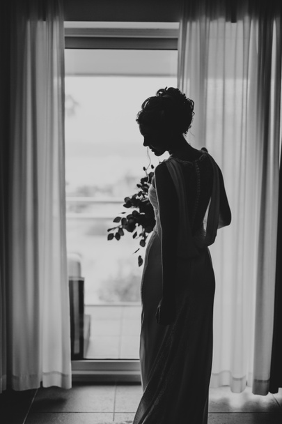 Hermosa novia en un vestido con un ramo de flores se encuentra, dando la vuelta y mirando al suelo en el dormitorio sobre el fondo de la ventana. Foto en blanco y negro - Foto, Imagen