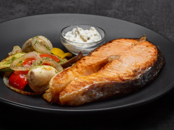 Roasted Salmon steak served with vegetables and creamy sauce on black plate. Stewed zucchini, bell pepper, onions near main dish. Fish with garnish on stone background - Photo, Image