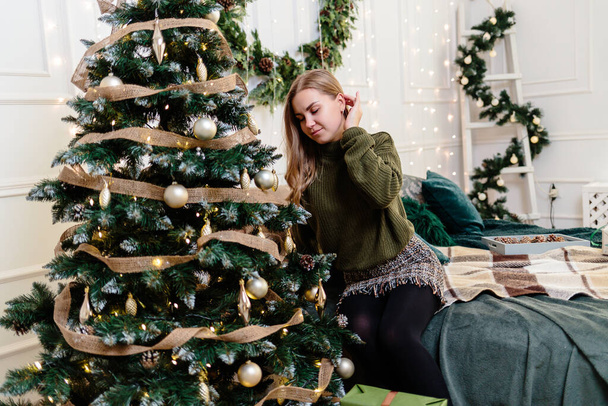 Μια όμορφη νεαρή γυναίκα με ξανθά μαλλιά κάθεται κοντά σε ένα χριστουγεννιάτικο δέντρο και διακοσμεί με παιχνίδια. Η Πρωτοχρονιά είναι σύντομα. Χριστουγεννιάτικη ατμόσφαιρα σε ένα άνετο σπίτι - Φωτογραφία, εικόνα