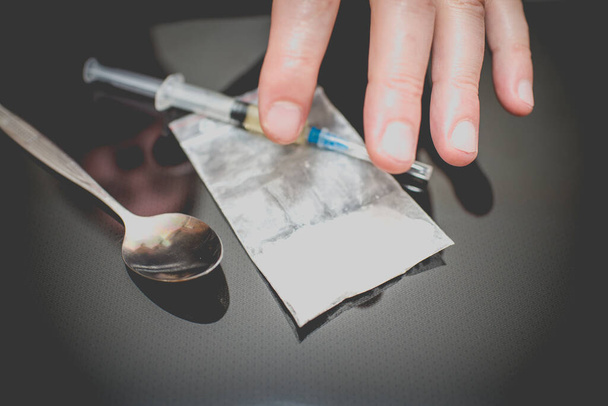 Close-up van verdovende drugsspuit, lepel, zak en cocaïne of heroïne op de tafel. De hand van de man op de achtergrond vervaagde.. - Foto, afbeelding