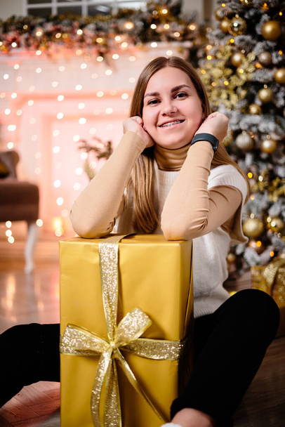 Eine Frau im weißen Pullover, ein warmer und gemütlicher Abend im weihnachtlichen Interieur, ein Weihnachtsbaum, geschmückt mit Lichtern, Geschenken, Spielzeug, einer Girlande. Festliches Wohnzimmer. Neues Jahr - Foto, Bild
