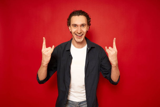 Happy overjoyed beautiful male with curly hair, shows cool sign love, says: Yeah, that wonderful Pretty young man gebaren indoor against red background copy space. Het begrip mens en lichaamstaal - Foto, afbeelding