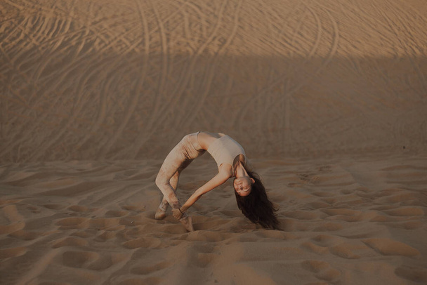 Krásná mladá brunetka v béžových šatech pózující na písečné pláži. Atraktivní žena šťastně tančí, pózuje na pouštním pozadí - Fotografie, Obrázek