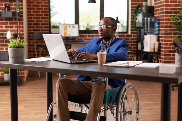 Επιχειρηματίας που κάθεται σε αναπηρικό καροτσάκι και εργάζεται σε φορητό υπολογιστή για να σχεδιάσει το έργο και τη στρατηγική στο γραφείο - Φωτογραφία, εικόνα