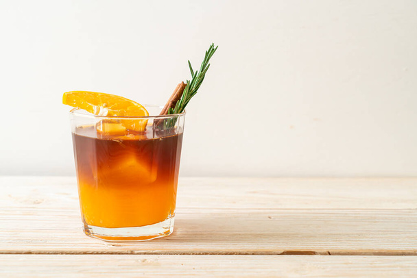 A glass of iced americano black coffee and layer of orange and lemon juice decorated with rosemary and cinnamon on wood background - Foto, immagini