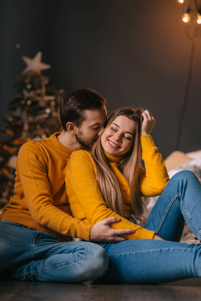 νεαρό όμορφο ζευγάρι κάθεται σε ένα όμορφο δωμάτιο στο κρεβάτι στο φόντο του χριστουγεννιάτικου δέντρου και χαμογελώντας. - Φωτογραφία, εικόνα