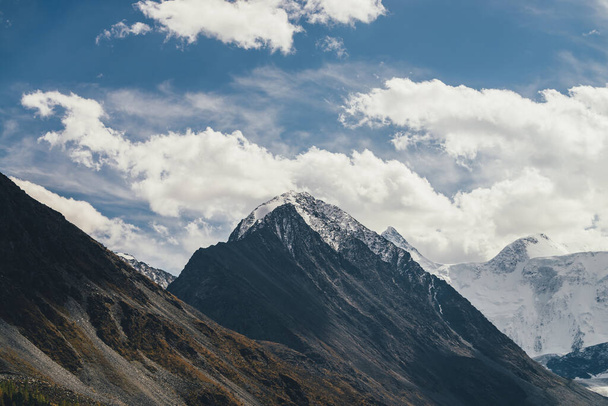 Paysage alpin atmosphérique avec silhouette de haute montagne avec neige sur le sommet culminant sous un ciel nuageux bleu. Paysage montagneux dramatique avec beau pic pointu enneigé et mur de montagne enneigé élevé - Photo, image