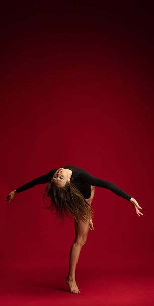 Studio shot of young flexible girl, female contemp dancer, ballerina in action isolated on dark red background. Art, beauty, inspiration concept. - Foto, Imagem